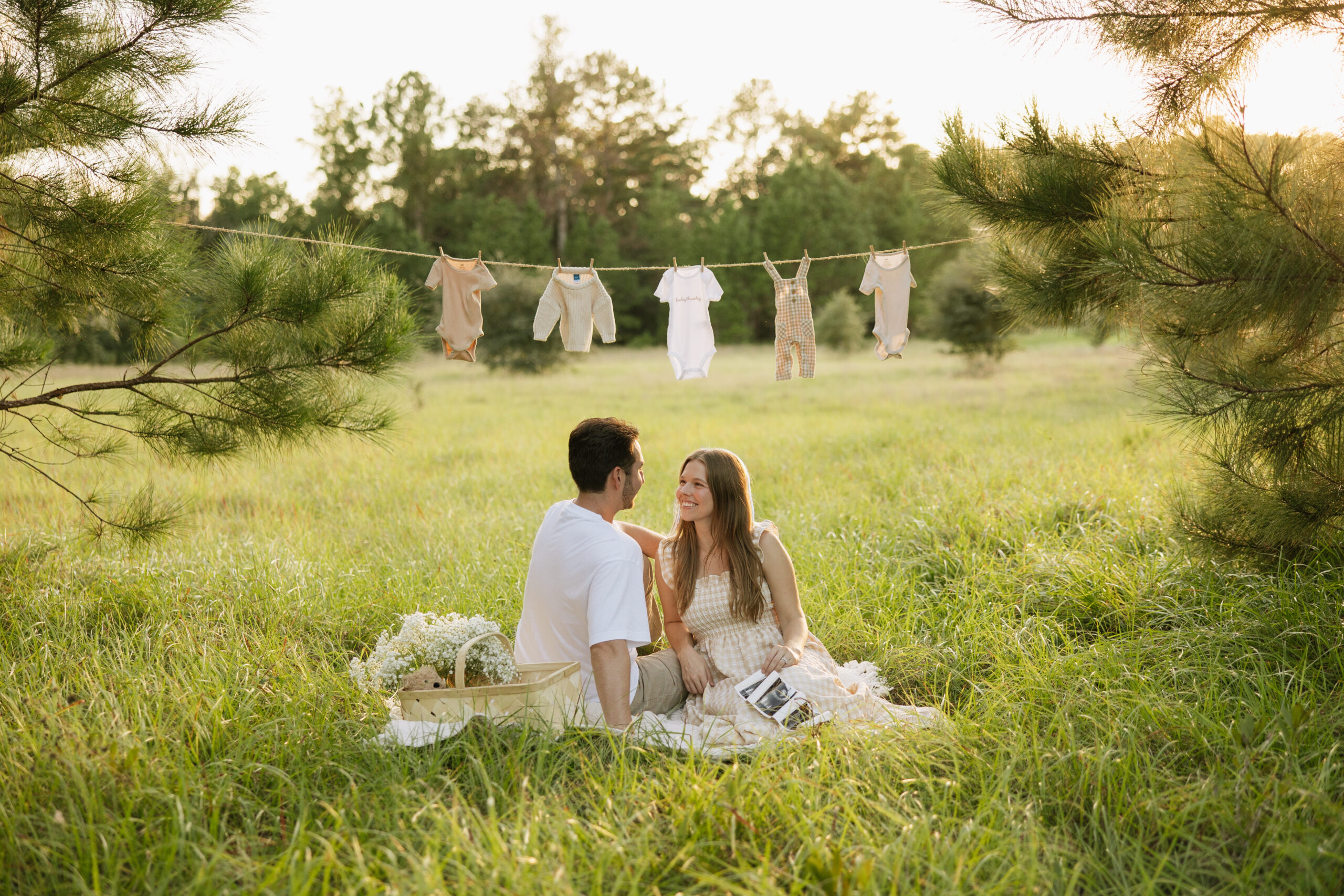 Couple sitting on a blanket with a clothes line of baby clothes. Holding ultrasound photos.